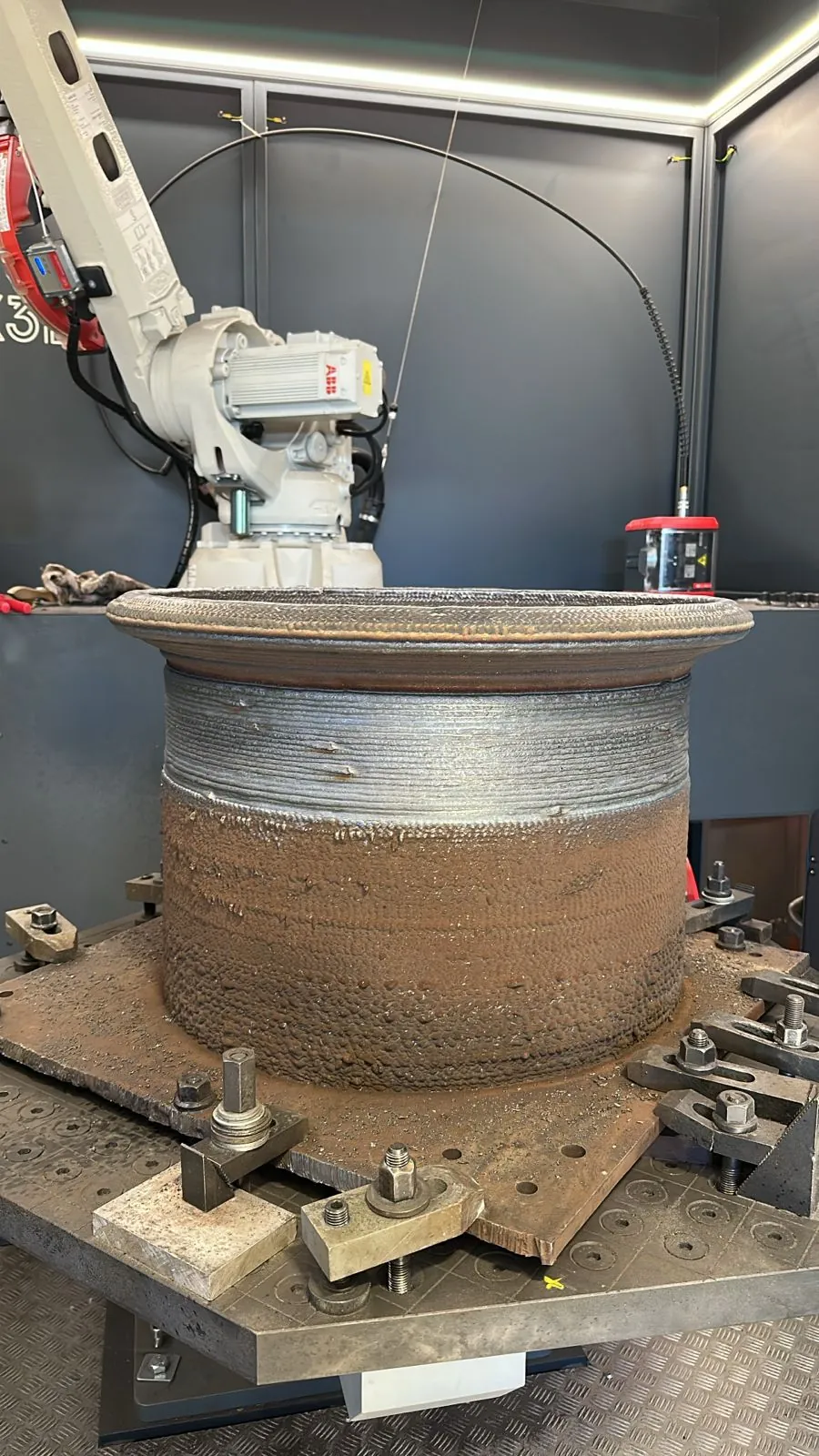 Peça cilíndrica de aço impressa em 3D por deposição a arco, com as camadas visíveis, diante do robô de solda no CDT MADA, em foto de divulgação da Belgo Arames