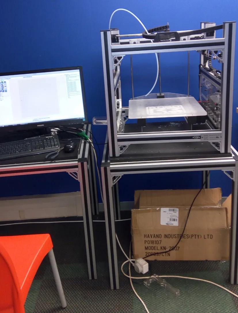 Impressora 3D de estrutura aberta, montada em perfil de aluminio, sobre bancada ao lado de um monitor — imagem ilustrativa, nao e a maquina citada na materia