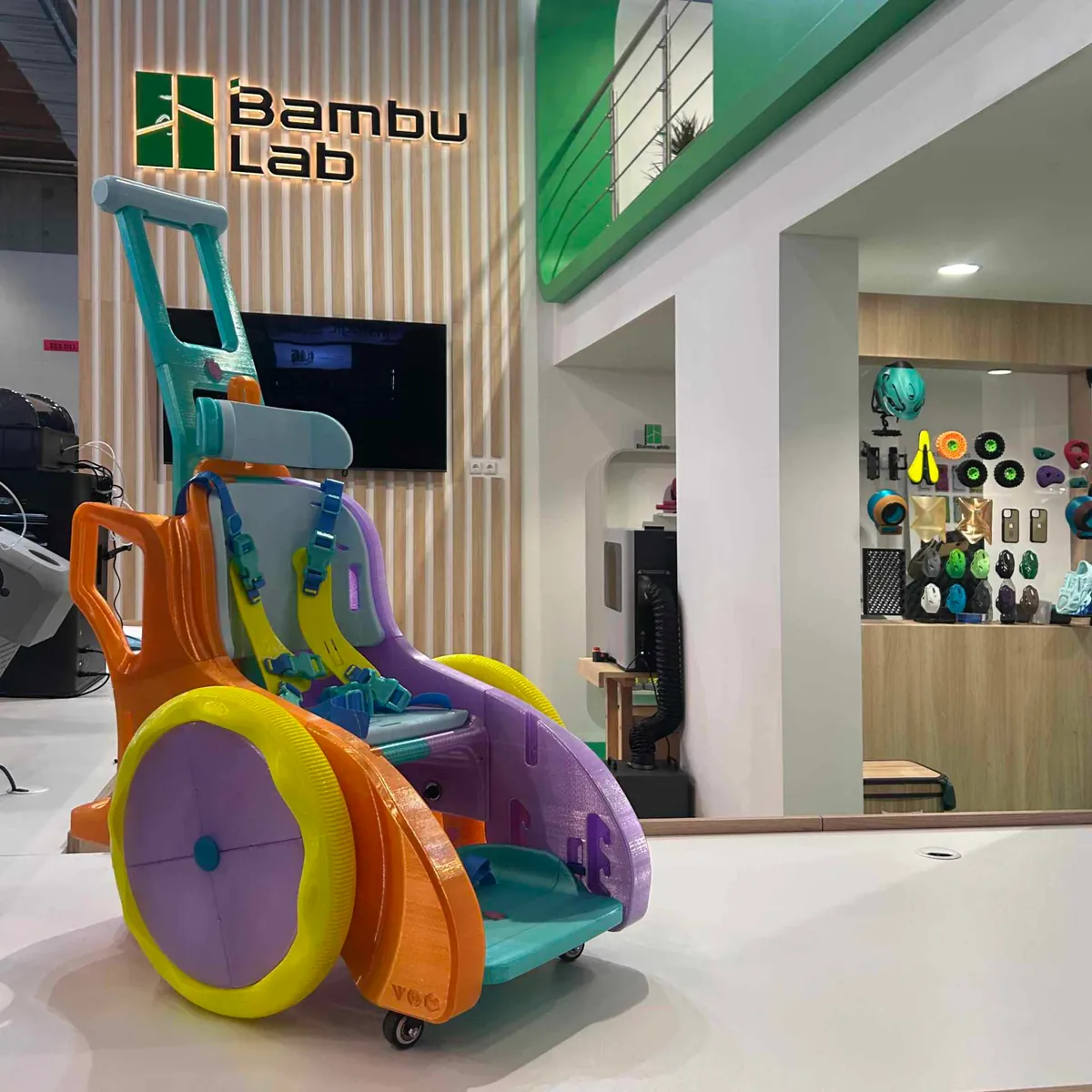 Andador infantil impresso em 3D em laranja, roxo, amarelo e azul, com assento acolchoado, cintos e rodas grandes, exposto num estande da Bambu Lab em feira