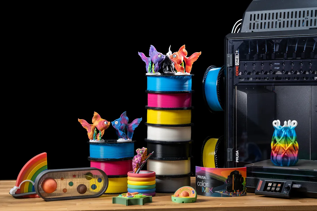 Rolos de Prusament PLA ColorMix em ciano, magenta, amarelo, branco e preto empilhados, com peixinhos impressos em tons mesclados e uma Prusa Core One L+ ao fundo