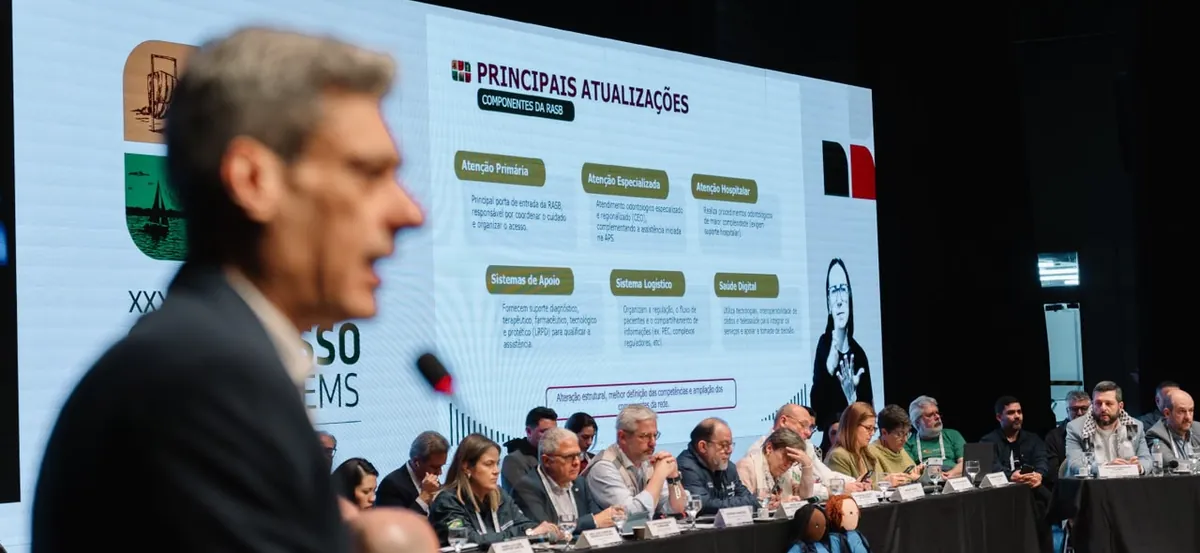 Reunião da Comissão Intergestores Tripartite em Porto Alegre: um homem de terno fala ao microfone em primeiro plano, desfocado, e ao fundo um telão mostra os componentes da Rede de Atenção à Saúde Bucal