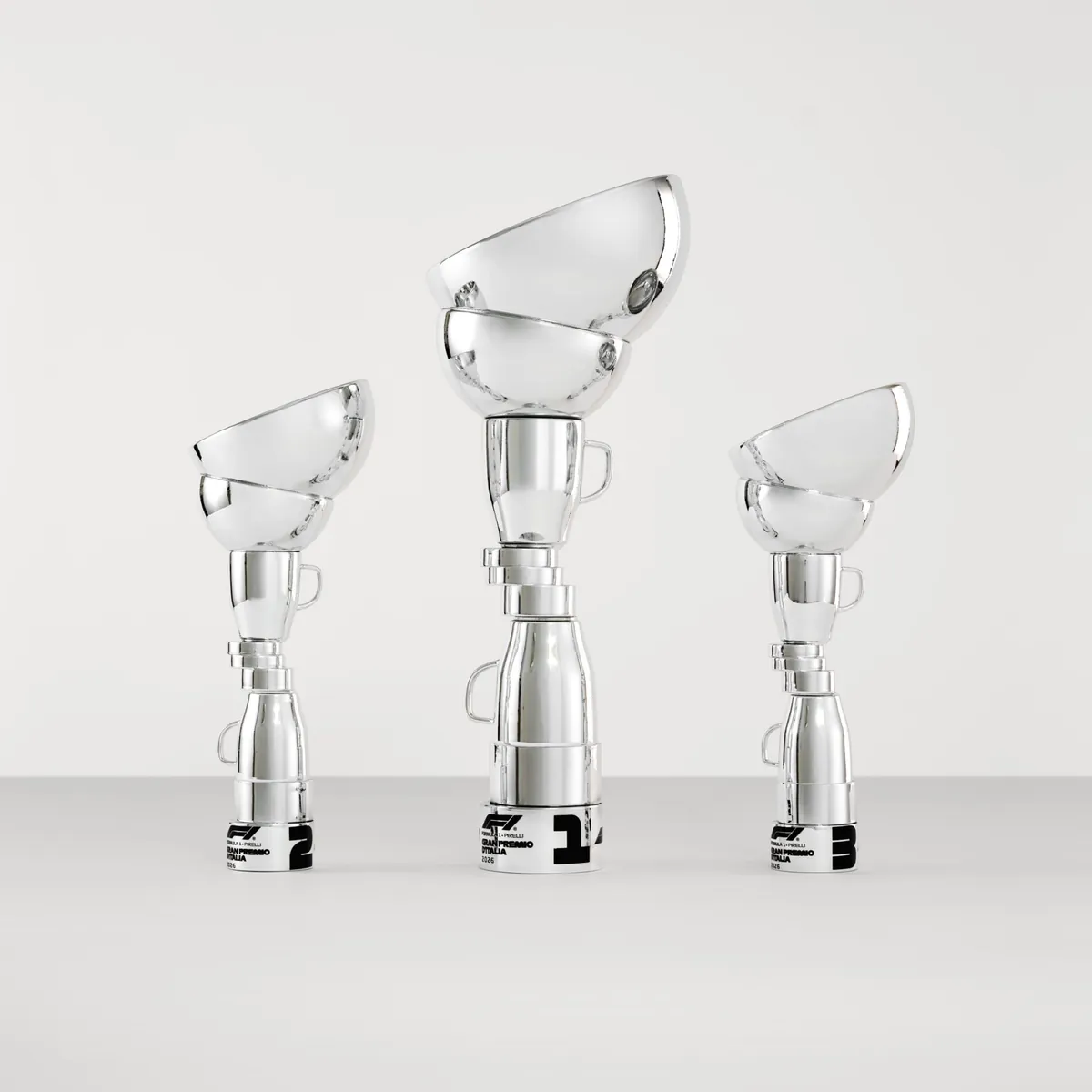 Três troféus prateados espelhados, formados por xícaras, tigelas e garrafas empilhadas numa torre instável, com as bases marcadas 1, 2 e 3 e o logo do GP da Itália 2026