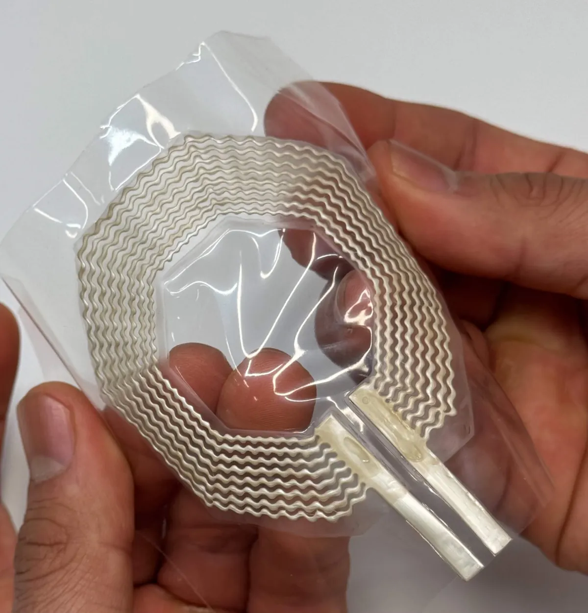 Mãos seguram uma bobina de ressonância magnética flexível e transparente, com trilhas onduladas de tinta de prata impressas em espiral sobre um filme elástico